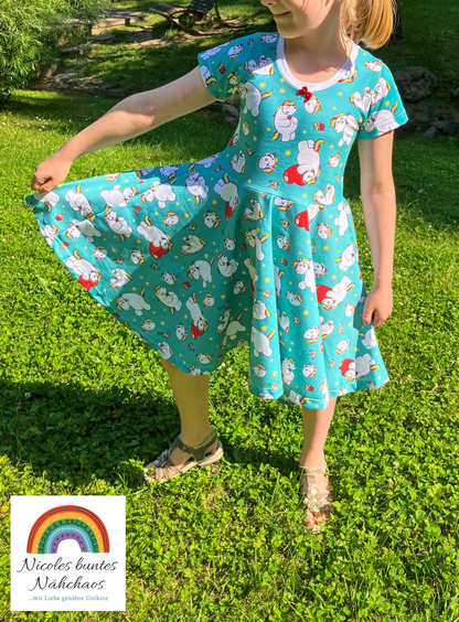 Mädchen trägt das Drehkleid "Marlene" mit dem Design "Pummel und Norbert Türkis" mit kurzen Ärmeln. Sie hält den Rock an der Seite hoch, um den Tellerrock zu präsentieren. Weißes Bündchen am Rundhalsausschnitt, kleine rote Schleife zur Verzierung.