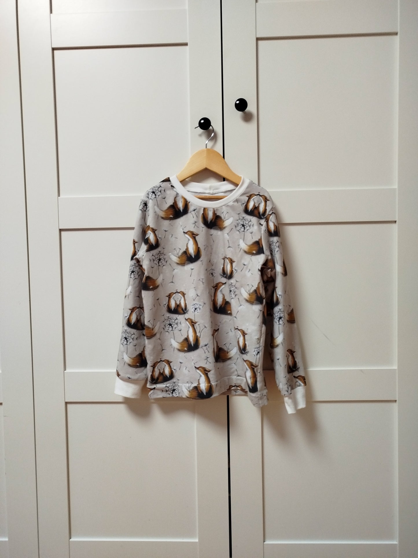 Beispielbild für einen Langarmsweater für Kinder mit langen Ärmeln und Bündchenabschluss an den Ärmeln und am Halsausschnitt. Rundhalsausschnitt. Hängend vor einer Schranktür. Farbe des Bündchens: Cremeweiß. Muster auf dem Sweater: Füchse mit Löwenzahnblumen.